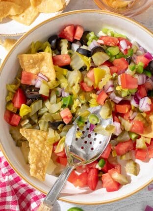 A white bowl filled with Pickle de Gallo made with dill pickles, tomato, onion, garlic. A spoon rests in the bowl.
