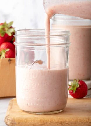 A clear glass mason jar half filled with Strawberry Banana Smoothie. Smoothie is being poured into the mason jar from a glass pitcher.