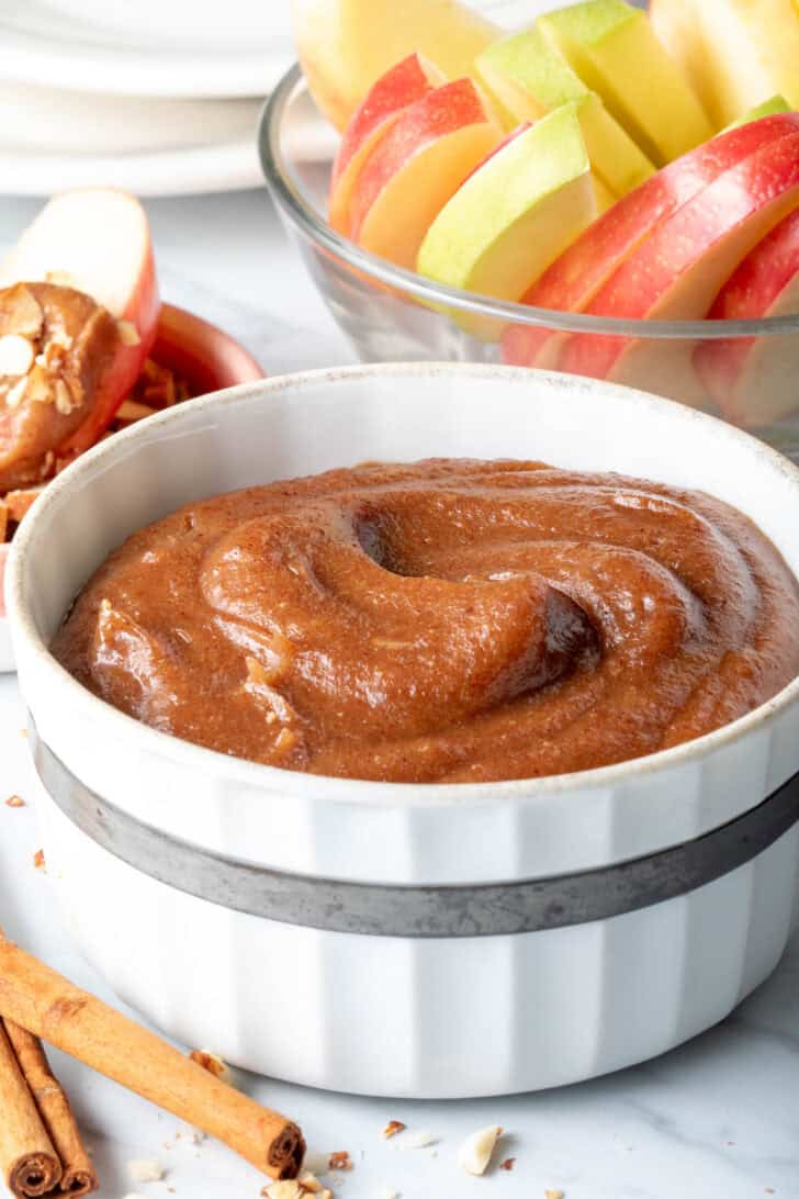 A small white bowl filled with Medjool Caramel Sauce (caramel apple dip). A clear glass bowl filled with apple slices sits next to the bowl.