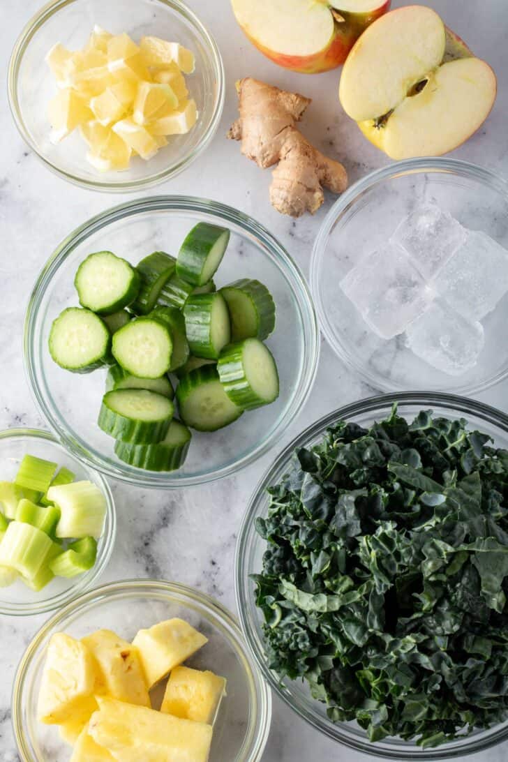 Clear glass bowls filled with chopped kale, cucumber, celery, pineapple lemon and apple.