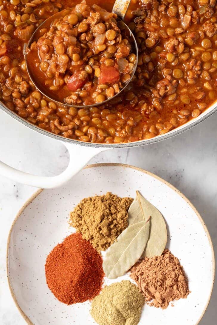 A small dish filled with the best homemade chili seasoning recipe ingredients sits next to a white pot of lentil chili.