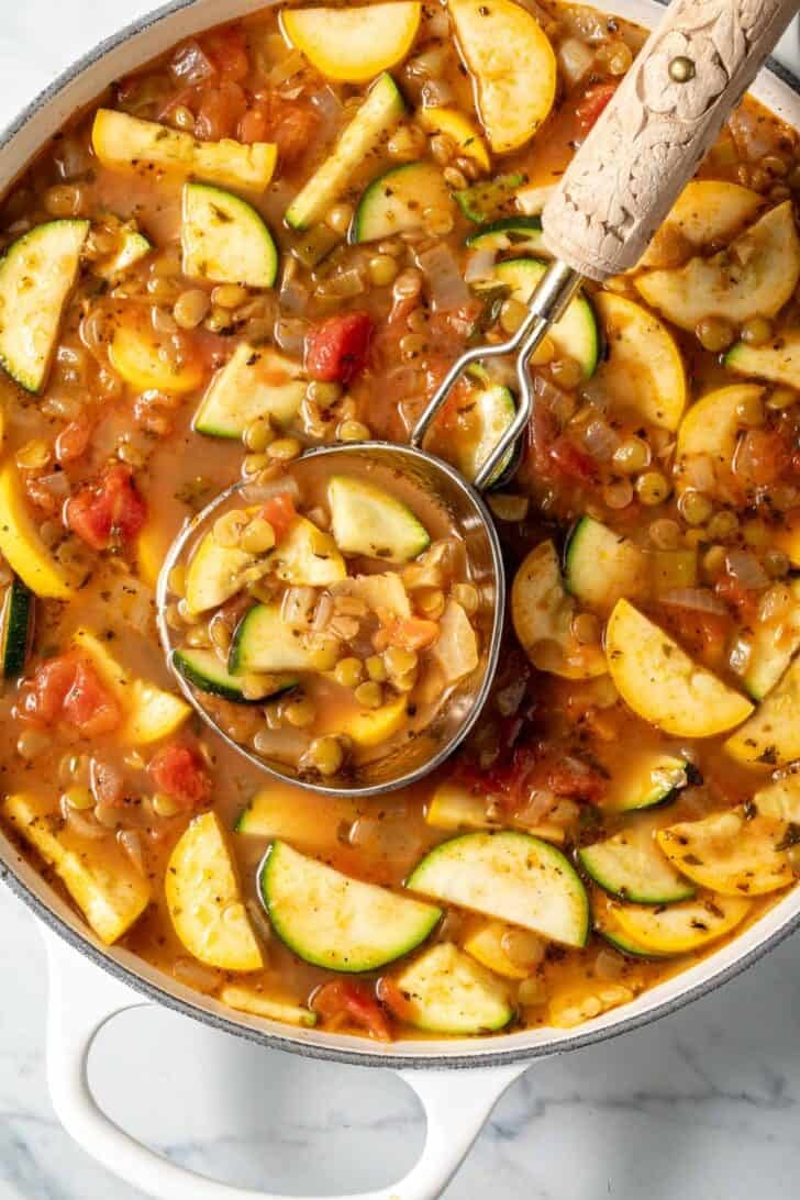 A white Dutch oven filled with vegetable lentil soup. A ladle rests in the pot.