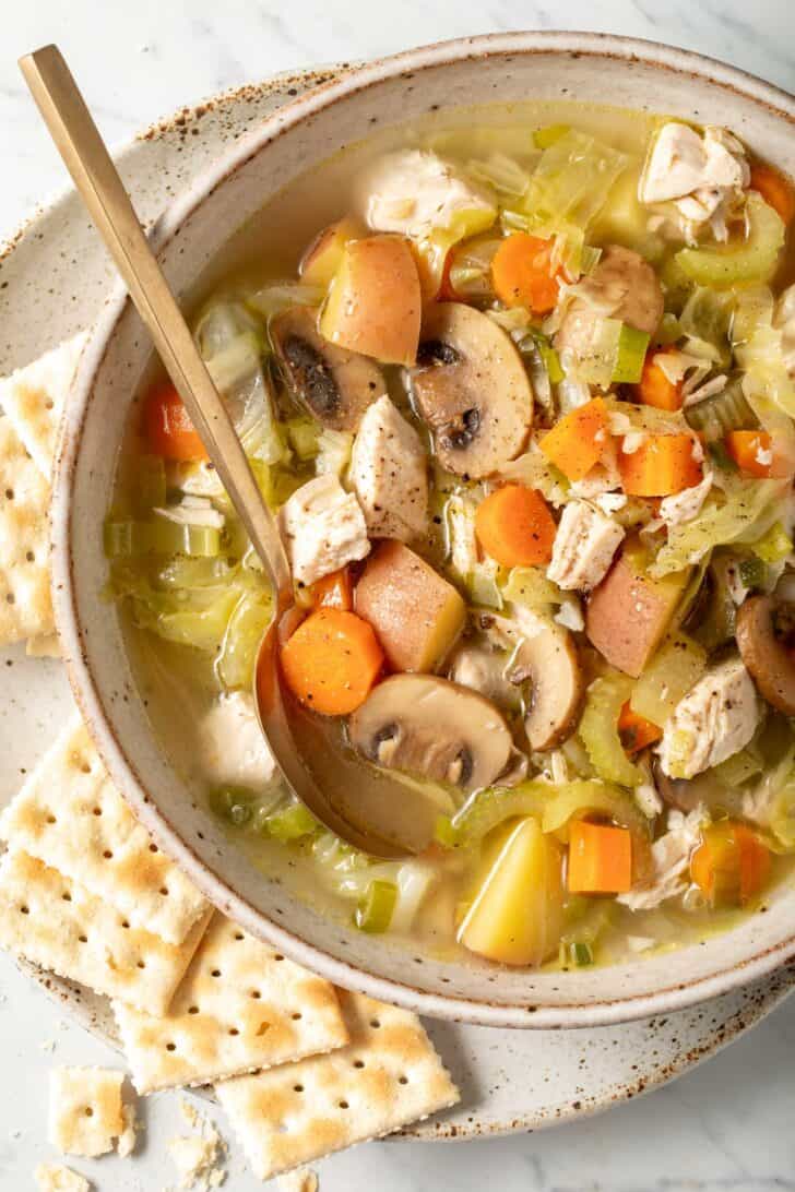 A bowl of cabbage soup with chicken and mushrooms in a soup bowl. Saltine crackers are scattered next to the bow.