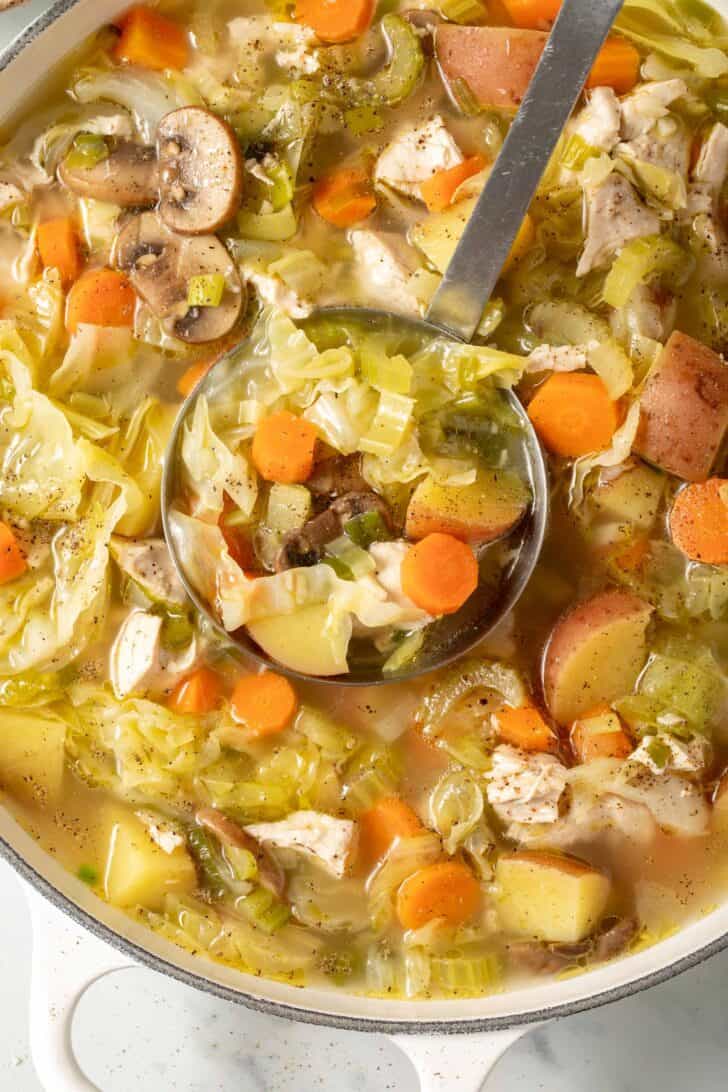 A white Dutch oven filled with chicken and cabbage soup. A ladle rests in the pot.