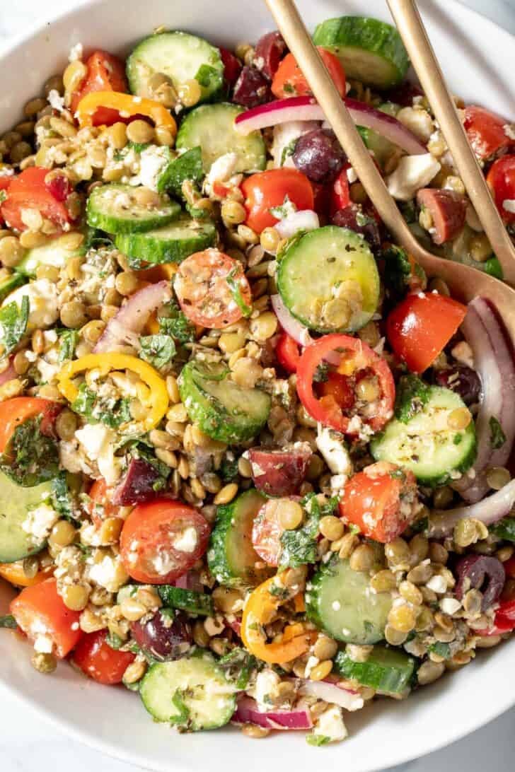 A white bowl filled with Greek Lentil Salad. Serving spoons rest in the bowl.