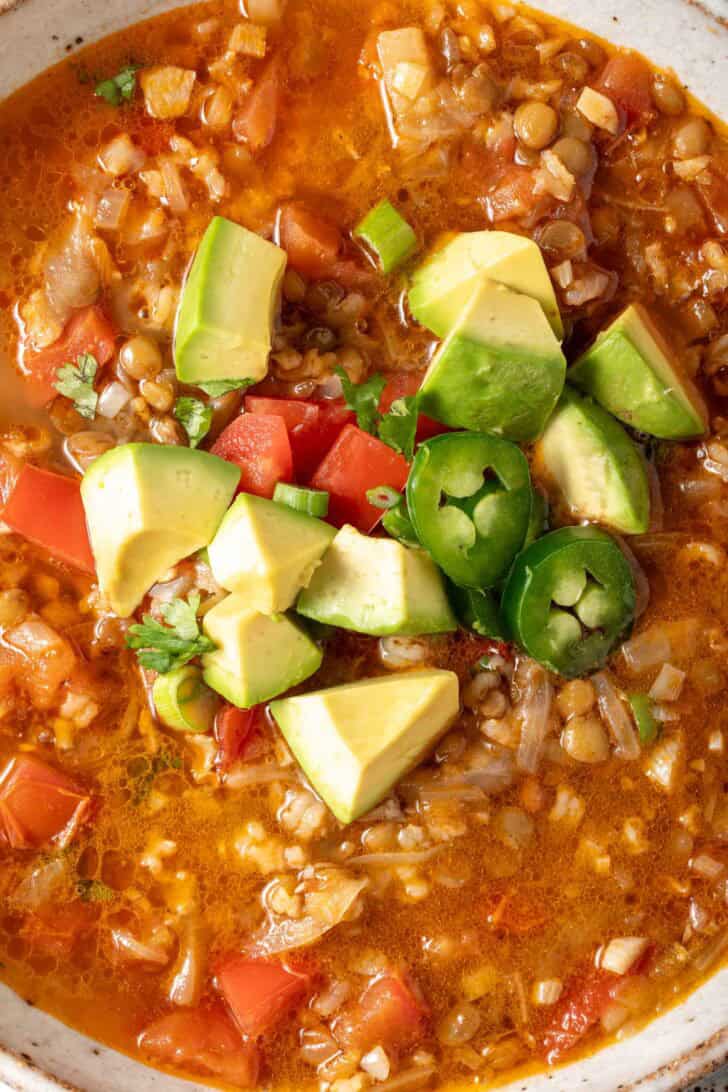 A bowl of lentil and rice soup garnished with chopped avocado, tomato, green onion and cilantro.