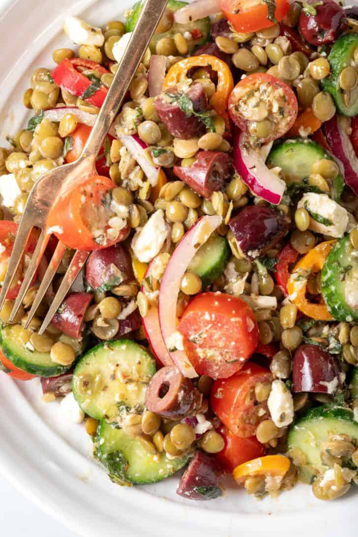 A white bowl filled with Greek Lentil Salad made with cooked lentils, sliced cucumber, onion, grape tomatoes, kalamata olives, sweet peppers and Feta cheese.