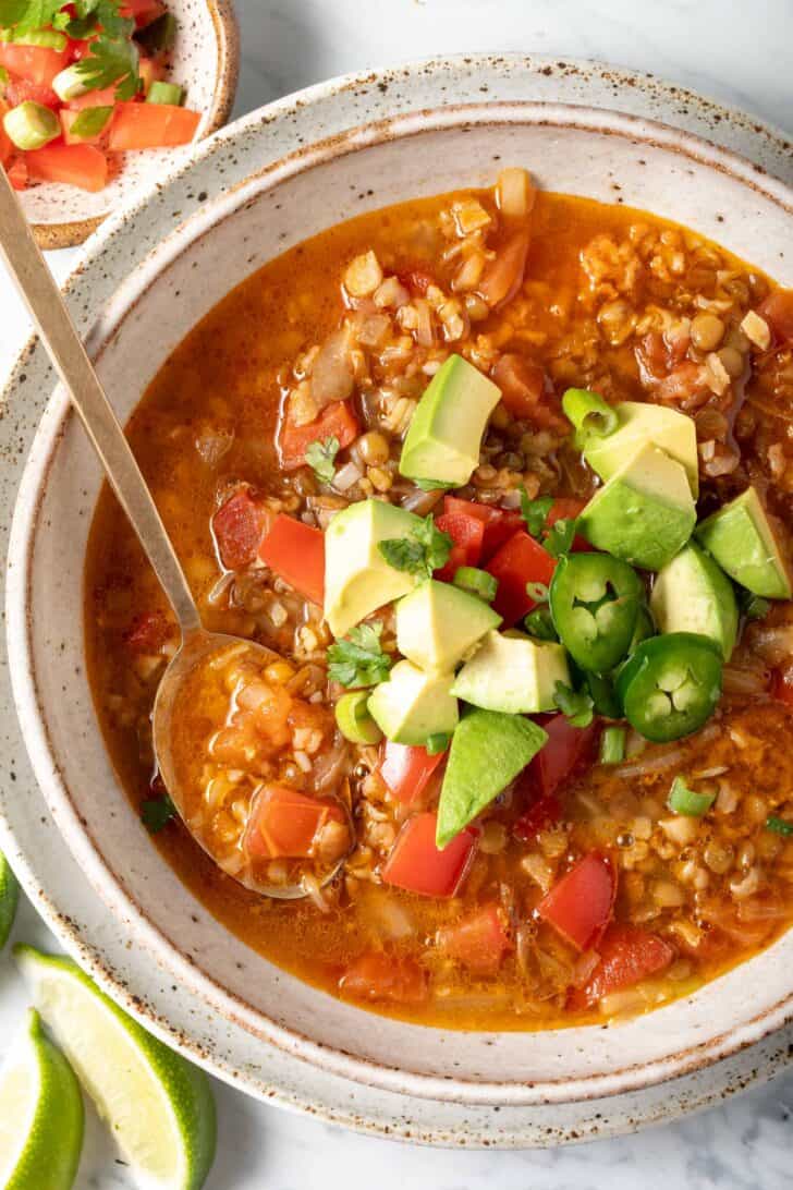 A bowl of lentil rice soup garnished with chopped avocado, tomato, green onion and cilantro.