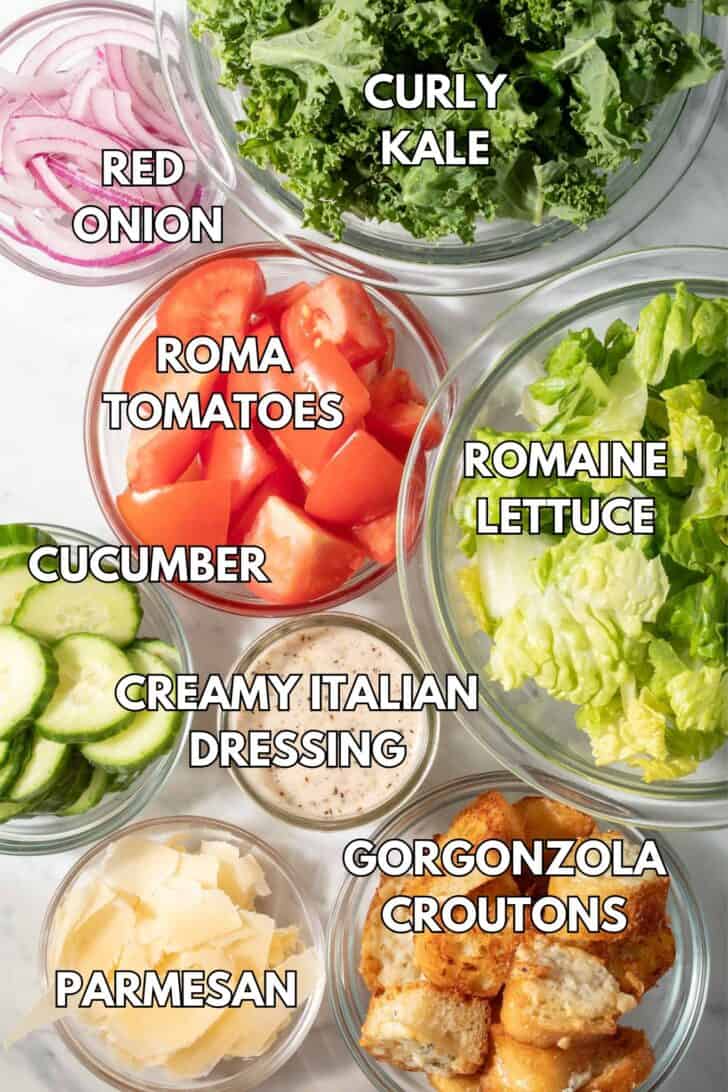 Clear glass bowls filled with lettuce, tomatoes, cucumber, grated cheese, croutons and sliced onion.