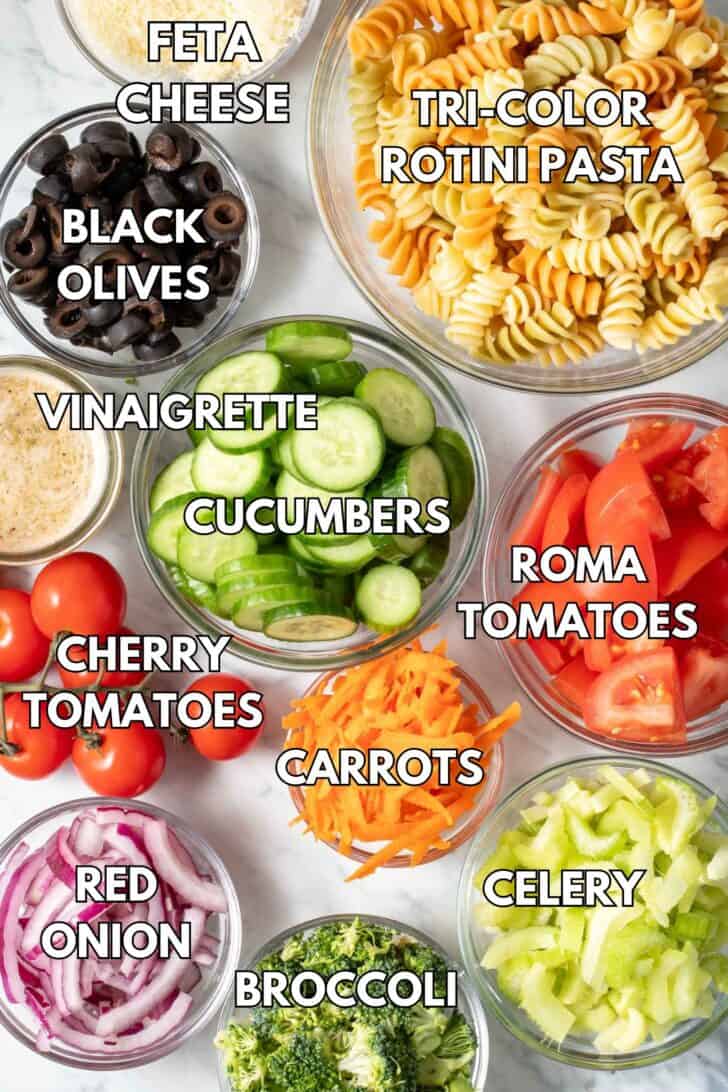 Clear glass bowls filled with pasta, cucumbers, tomatoes, celery, broccoli, carrots, feta cheese and dressing.
