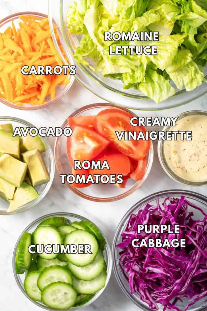 Clear glass bowls filled with lettuce, cabbage, tomatoes, carrots, cucumber and avocado.