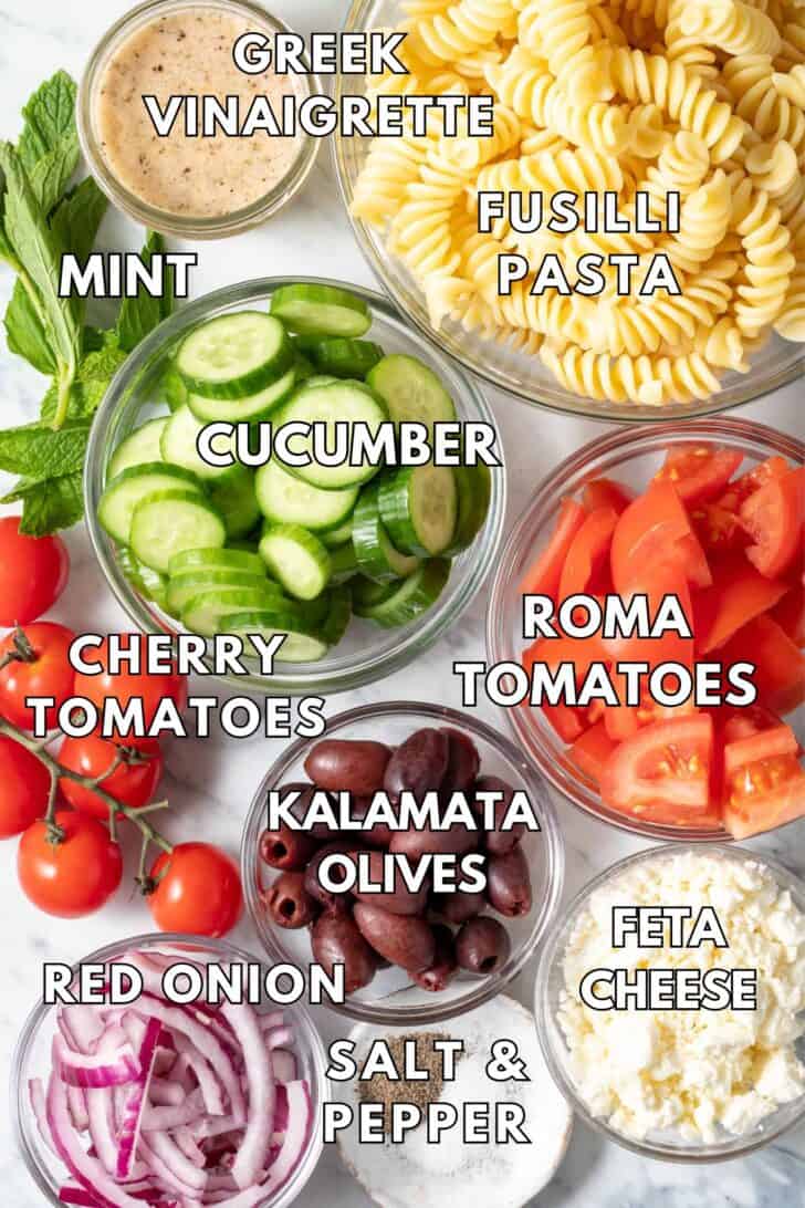 Glass bowls filled with pasta, tomatoes, cucumbers, red onion, feta cheese and kalamata olives.