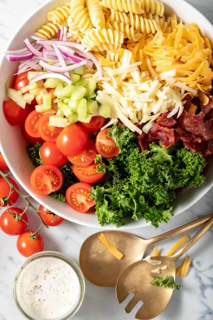 A white bowl filled with chopped curly kale, cherry tomatoes, celery, red onion, cheese and pasta noodles.