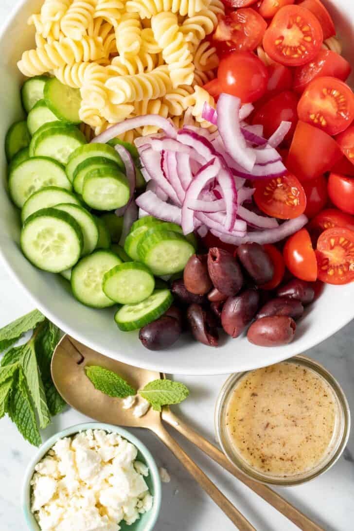 A white bowl filled with sliced cucumbers, tomatoes, olives, onion and noodles for a Greek Pasta Salad recipe.