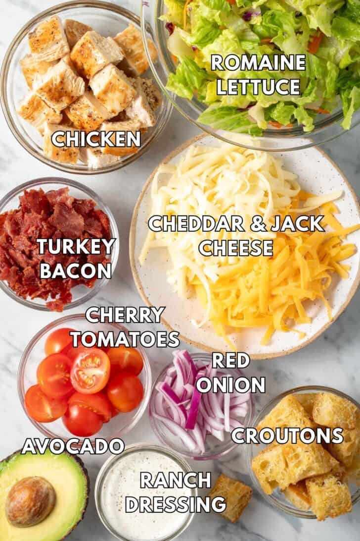 Clear glass bowls filled with chopped lettuce, grated cheese, cherry tomatoes, red onion and croutons.