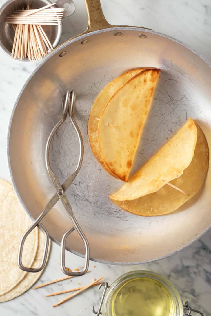 A skillet with two hard taco shells in it with a pair of tongs.