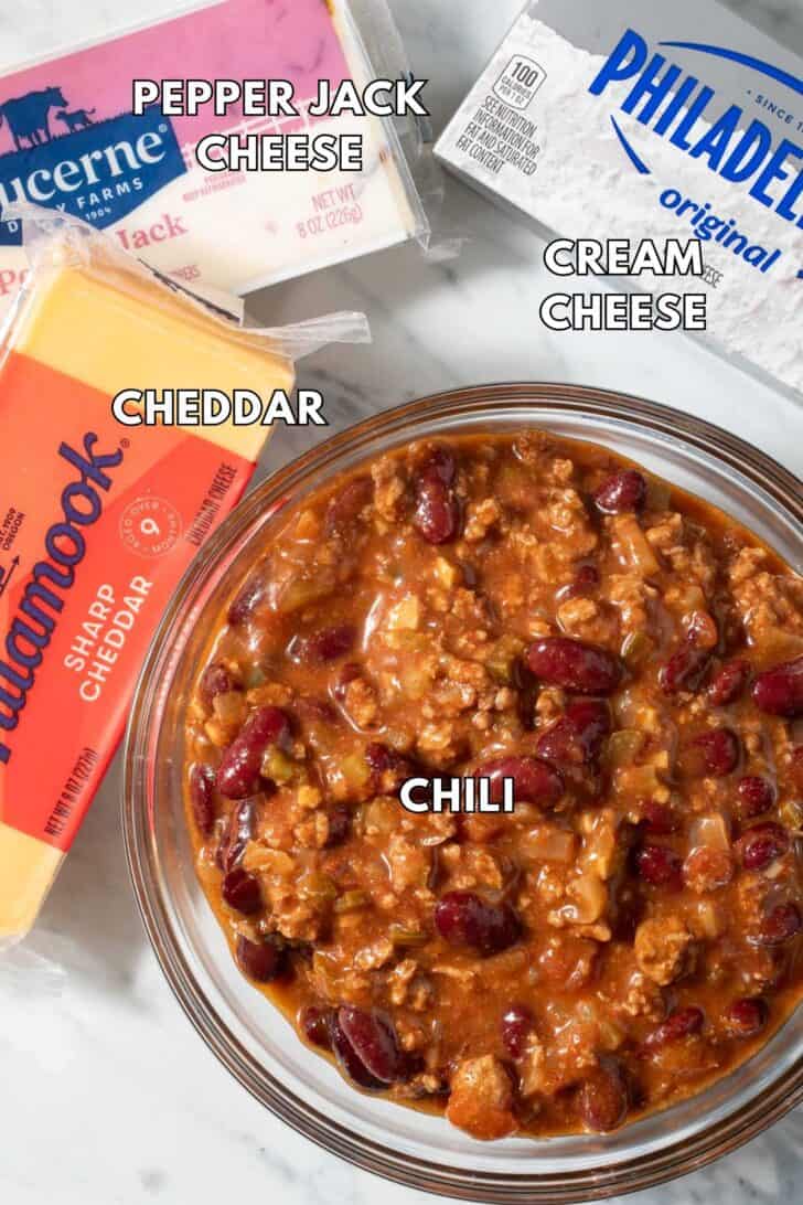 Chili in a glass bowl, with a package of sharp cheddar cheese, pepper Jack cheese and cream cheese.