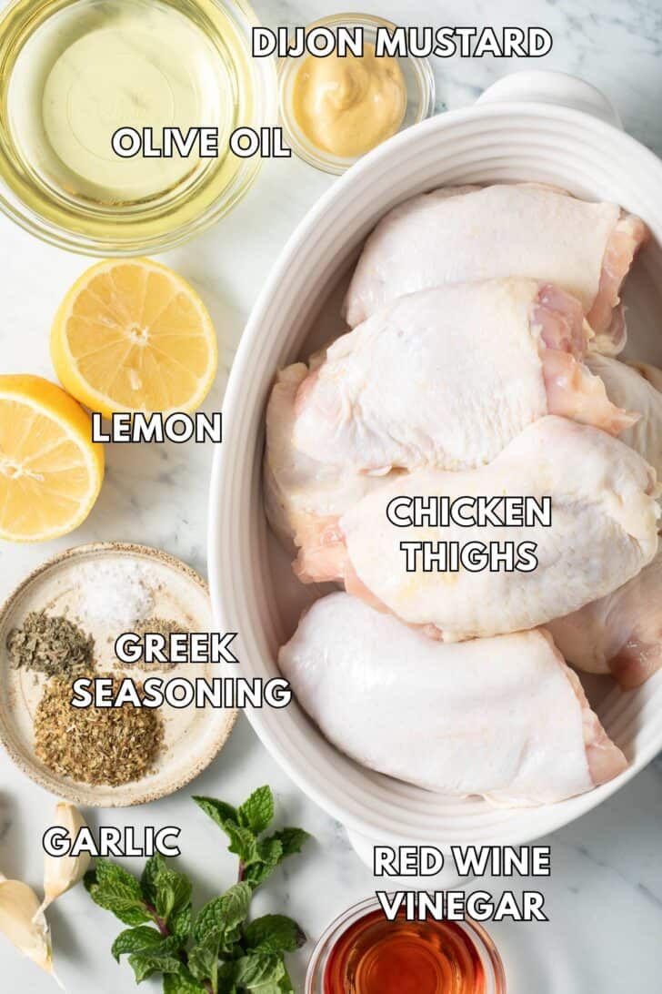 Greek Chicken Recipe Raw chicken thighs in a bowl with seasonings, lemon and olive oil next to it.
