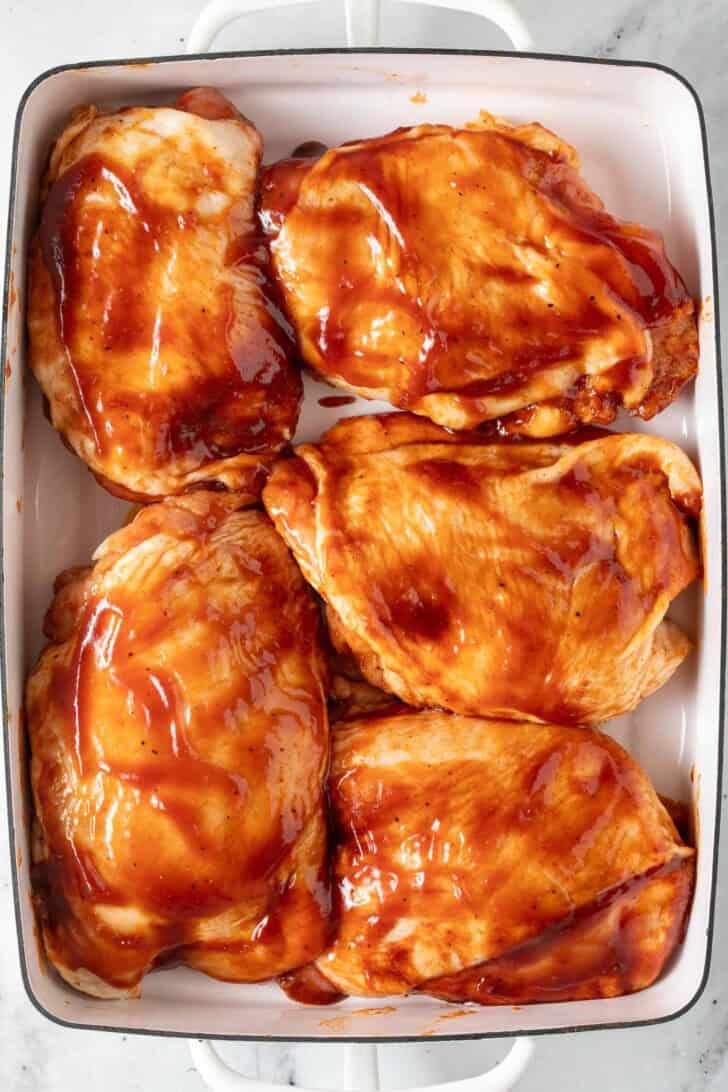 BBQ Chicken Thighs In oven Chicken Thighs brushed with barbecue sauce in a white baking dish.