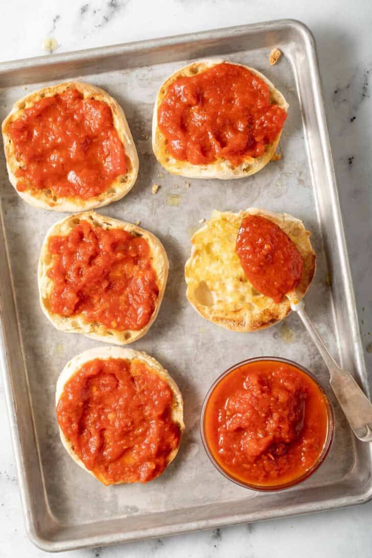 How to Make English Muffin Pizzas Toasted English muffins with marinara sauce on a baking sheet.