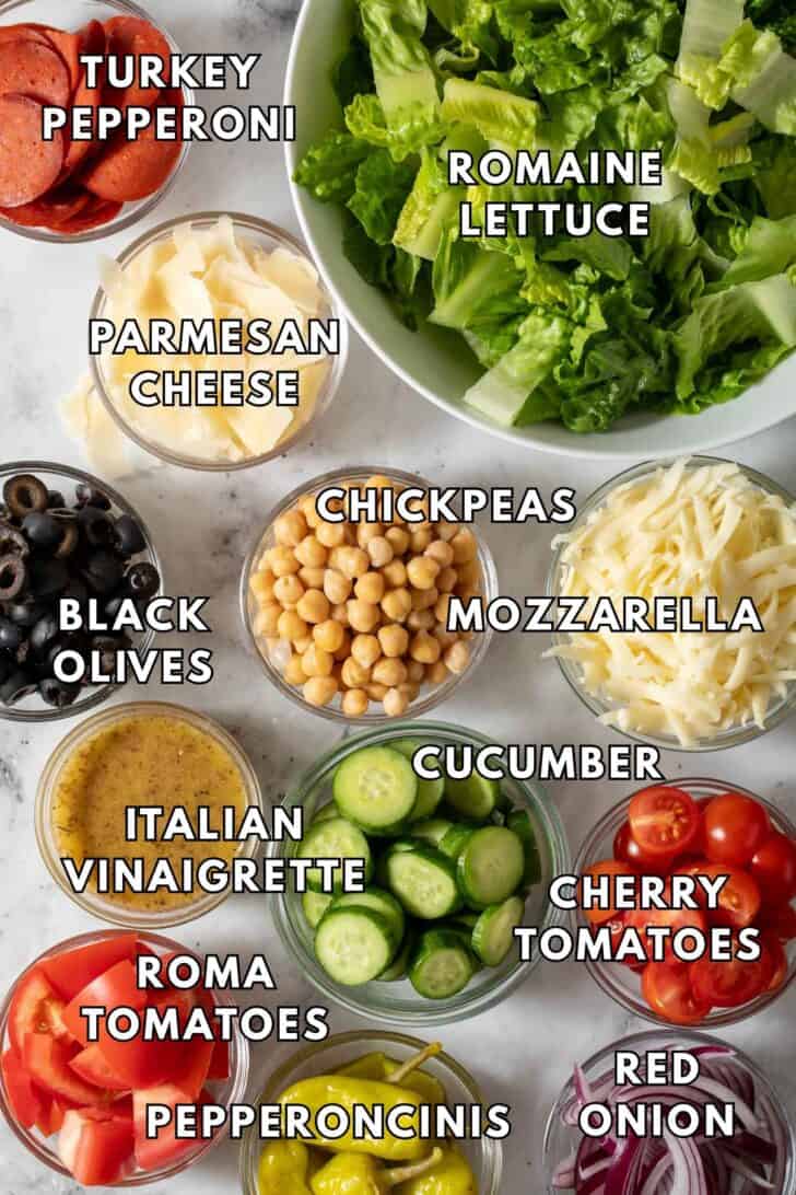 Pizza Salad Recipe Clear glass bowls filled with grated cheese, chickpeas, sliced cucumber, sliced tomatoes and red onions for a Pizza Salad recipe.