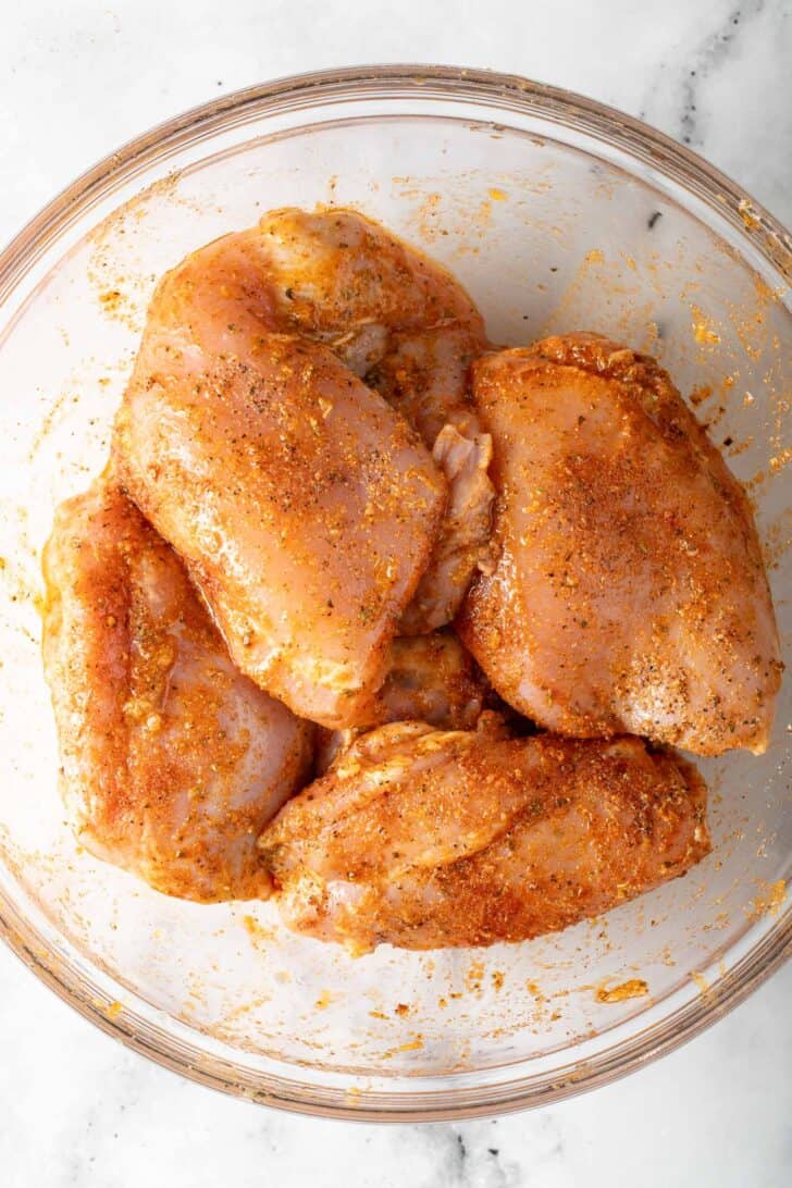 Boneless Skinless Chicken Thighs Raw boneless skinless chicken thighs tossed in seasoning in a glass bowl.