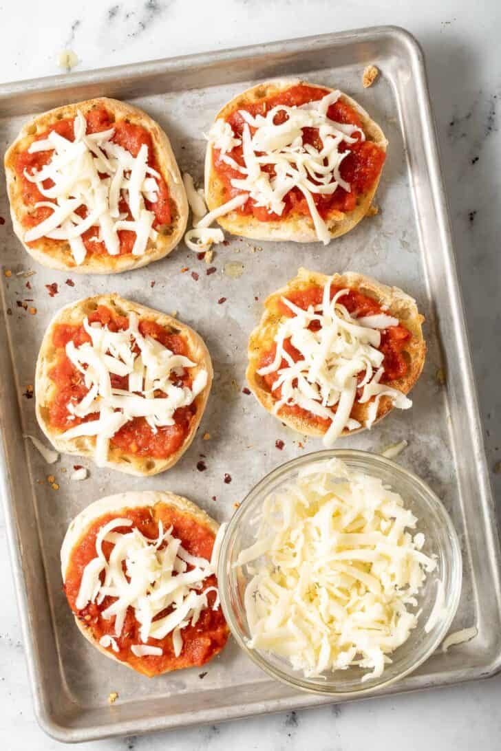 English Muffin Pizzas Recipe Toasted English muffins with marinara sauce and grated cheese on a baking sheet.