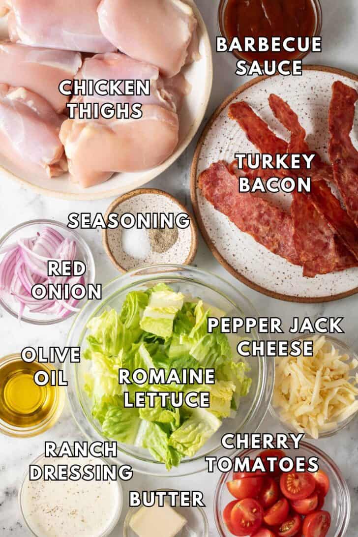 BBQ Chicken Salad Recipe Glass bowls filled with chopped lettuce, grated cheese, cherry tomatoes next to a plate of cooked bacon and chicken thighs.