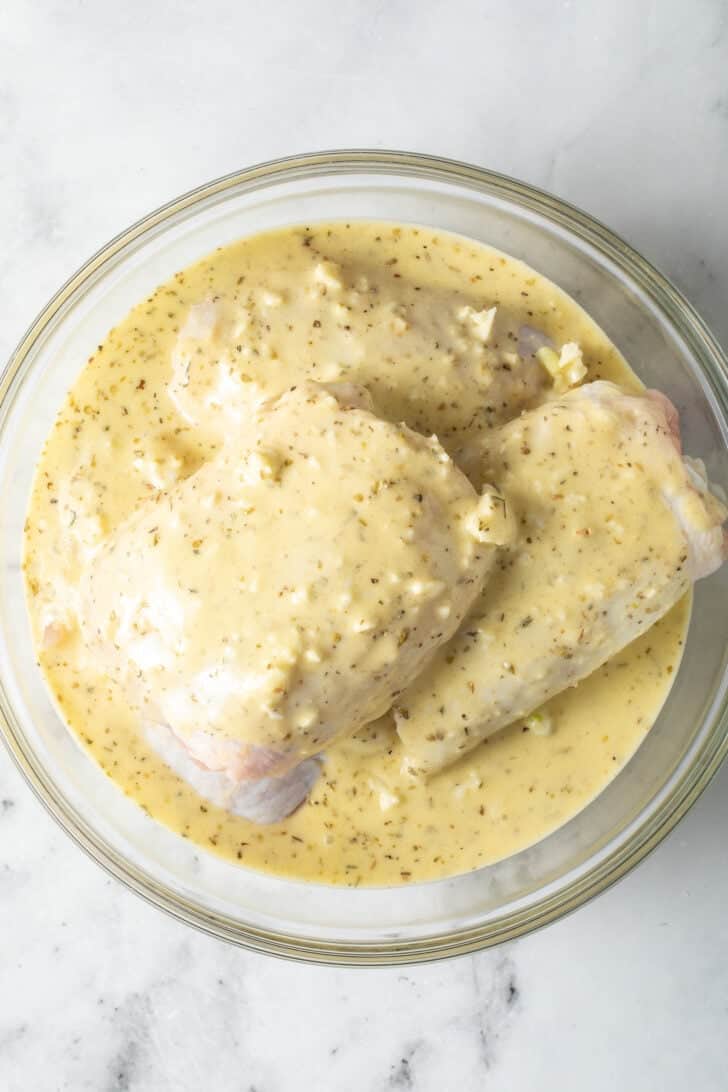 Greek Lemon Chicken Marinade Chicken thighs in a clear glass bowl marinating in Greek Lemon Chicken Marinade.