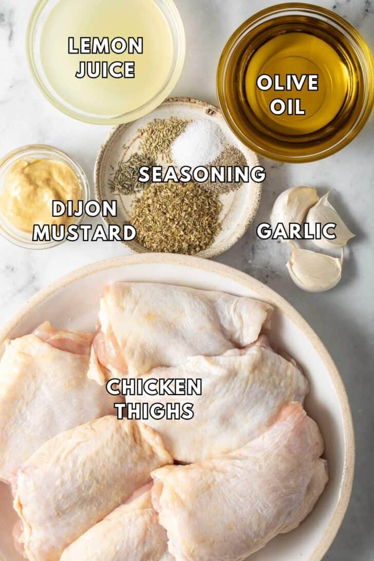 Greek Lemon Chicken Recipe A plate with chicken thighs next to clear glass bowls of lemon juice, olive oil, Dijon mustard and seasoning for a Greek Lemon Chicken recipe.