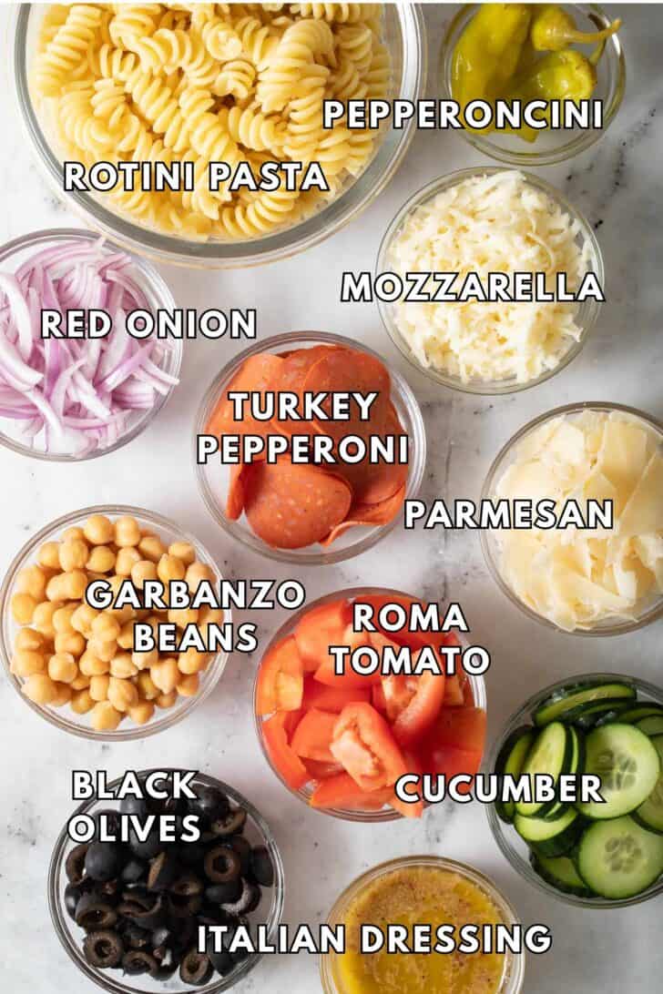 Rotini Pasta Salad Recipe Small glass bowls filled with cooked pasta noodles, grated mozzarella, garbanzo beans, and cucumber slices for a rotini pasta salad.