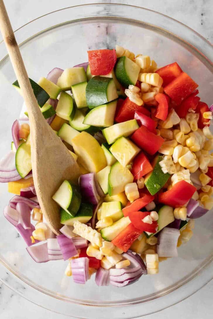 Summer Vegetables Recipe Chopped vegetables in a clear glass bowl with a wooden spoon.