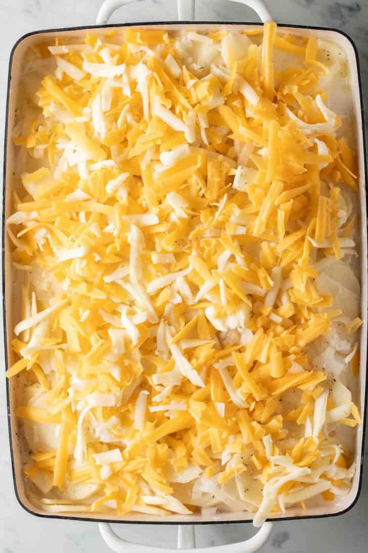 How to Make Au Gratin Potatoes Raw sliced potatoes and onions with grated cheese in a white baking dish.