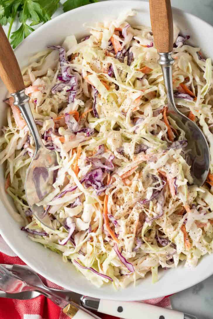 Simple Coleslaw Recipe Creamy coleslaw in a white bowl with serving spoons.