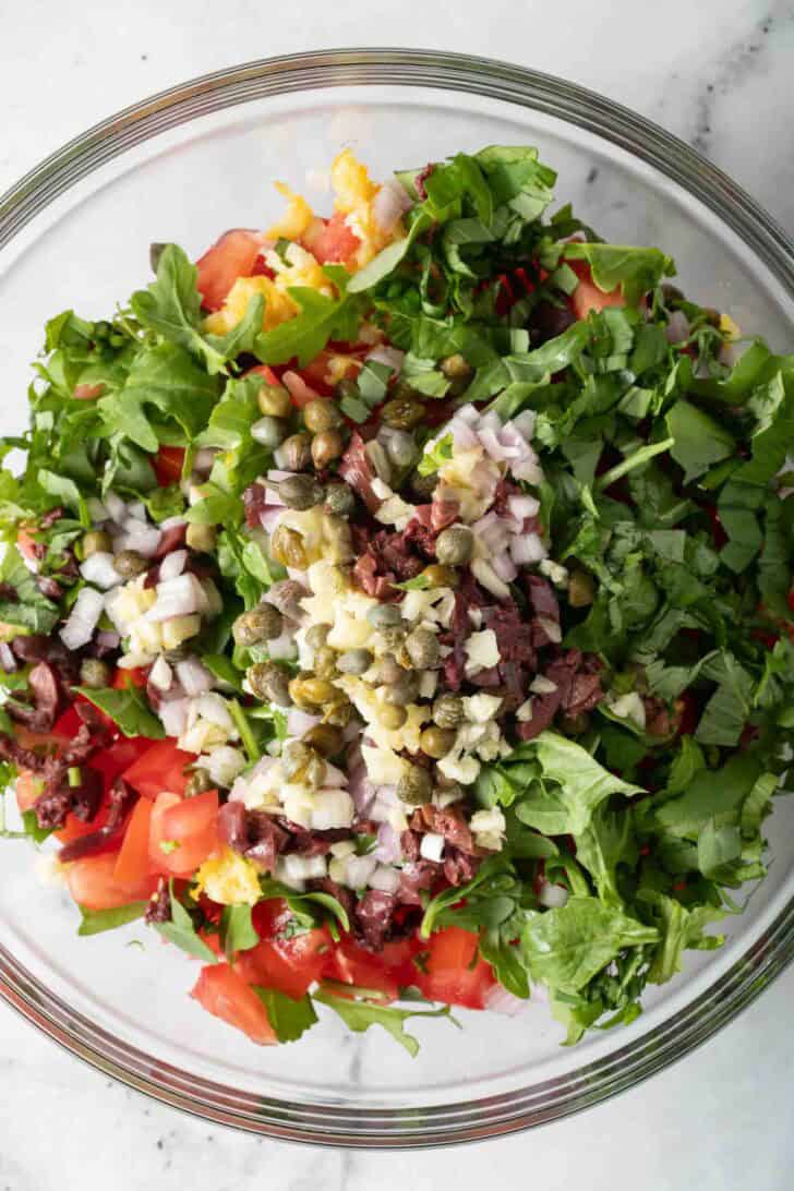 Roma Tomatoes For Bruschetta Clear glass bowl filled with chopped arugula, kalamata olives, capers, shallot and Roma tomatoes for bruschetta recipe.