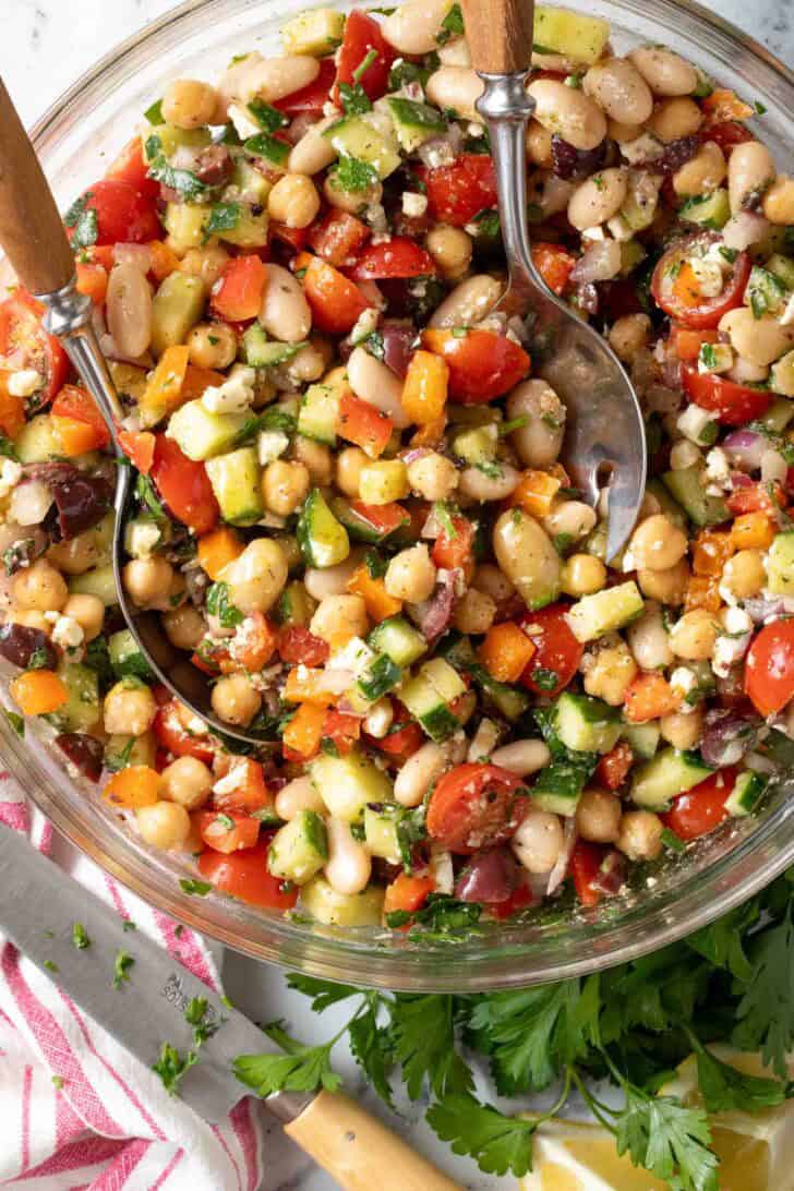 Greek Dense Bean Salad Greek Dense Bean Salad in a clear glass bowl with serving spoons.