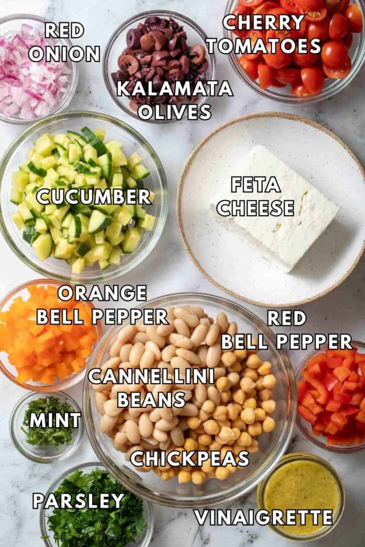 Greek Dense Bean Salad Recipe Clear glass bowls filled with chickpeas, cannellini beans, cherry tomatoes, onion, cucumber and bell pepper for a Greek Dense Bean Salad Recipe.