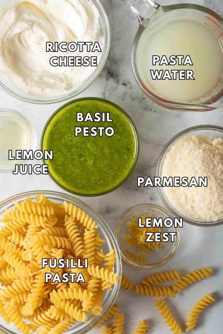 Creamy Pesto Pasta Recipe Clear glass bowls filled with basil pesto, grated parmesan cheese, lemon juice, ricotta cheese and fusilli pasta for a Creamy Pesto Pasta Recipe.