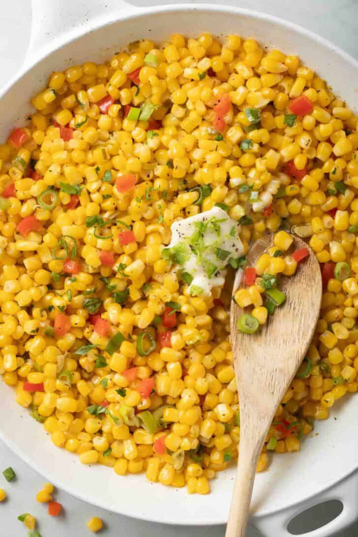Skillet Corn Recipe Skillet Corn in a white skillet with a wooden spoon.