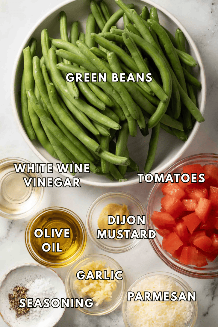 Green Bean Salad Recipe Fresh green beans in a white bowl next to clear glass bowls of chopped tomatoes and parmesan cheese for a Green Bean Salad recipe.