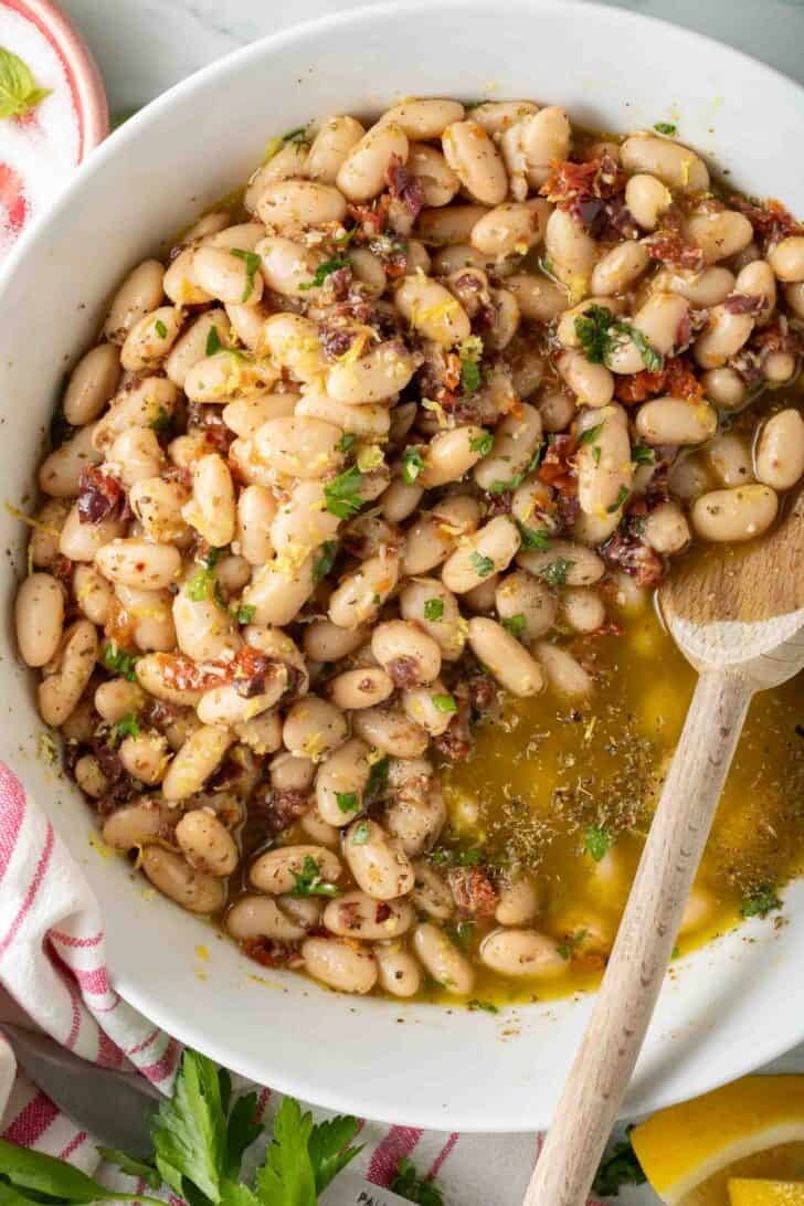 Marinated White Beans Marinated White beans in a white bowl with a wooden spoon.