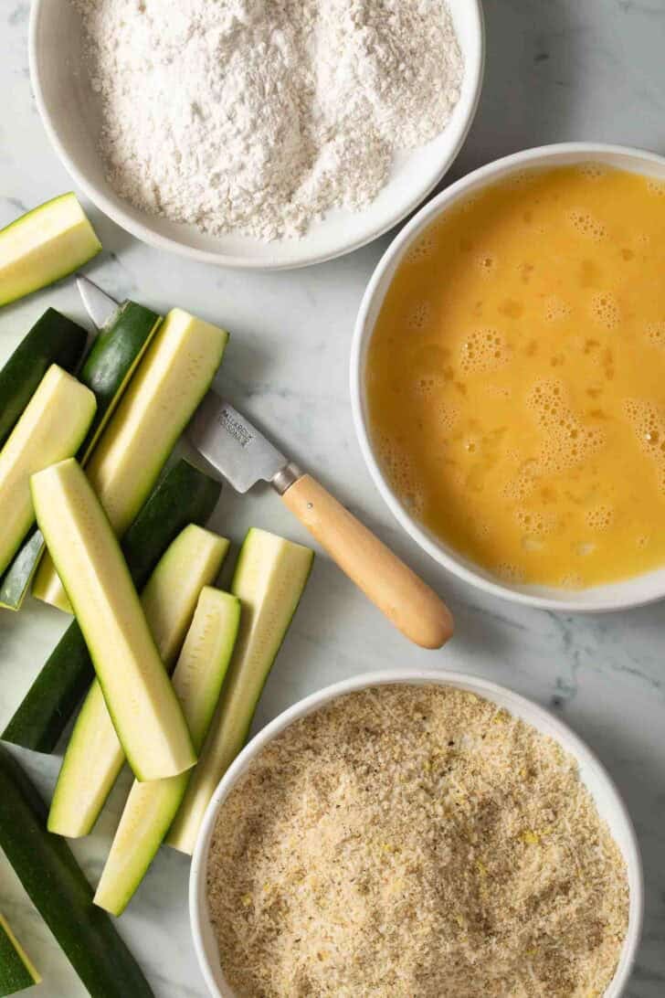 Zucchini Sticks Recipe Zucchini sticks next to bowls of flour, egg, and bread crumbs for zucchini fries recipe.