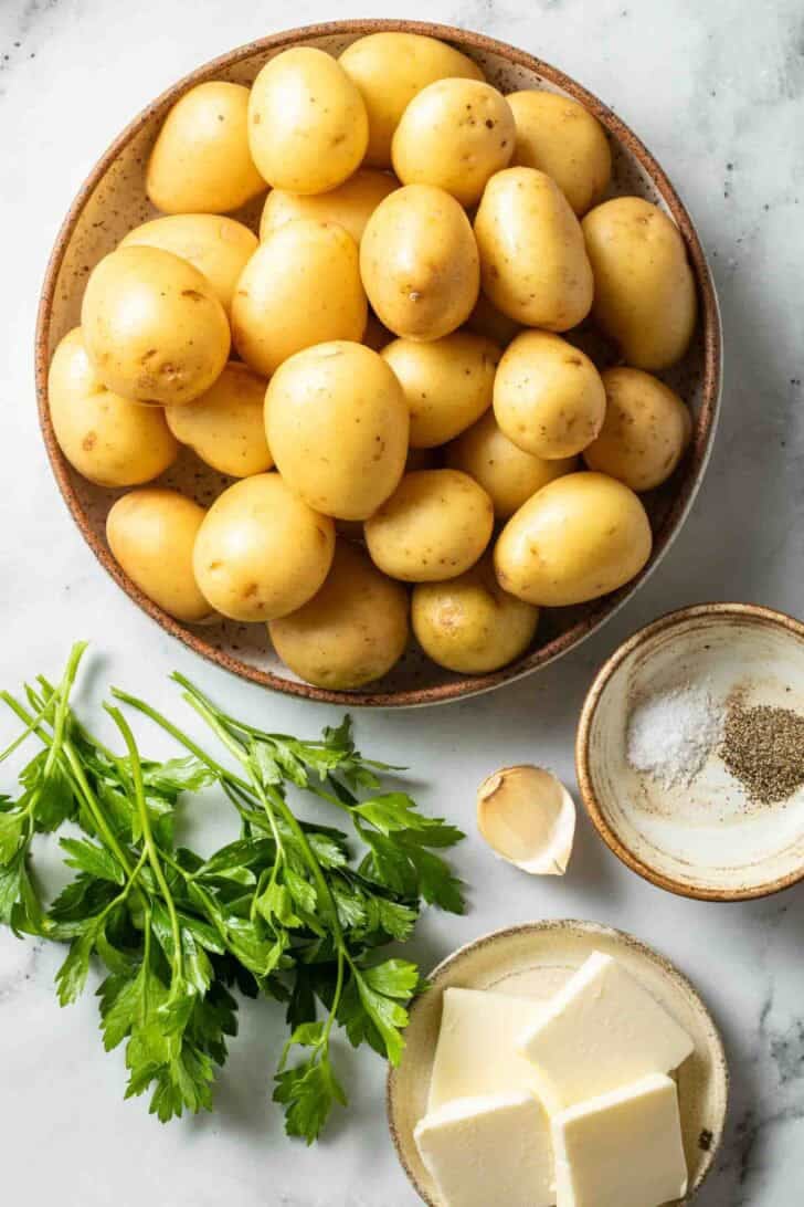 Boiled Potatoes Recipe Baby Yukon gold potatoes in a bowl next to sliced butter, fresh parsley, garlic and seasoning.