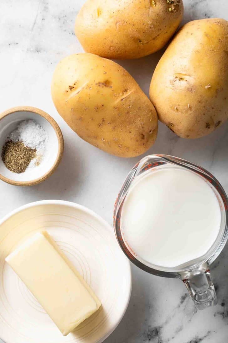 Yukon Gold Mashed Potatoes Recipe Yukon gold potatoes next to a stick of butter, a cup of milk and salt and black pepper.