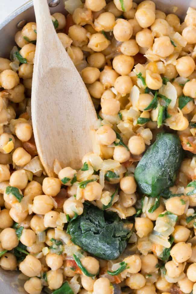 An aluminum skillet filled with chickpeas and spinach. A wooden spoon rests in the pan. 