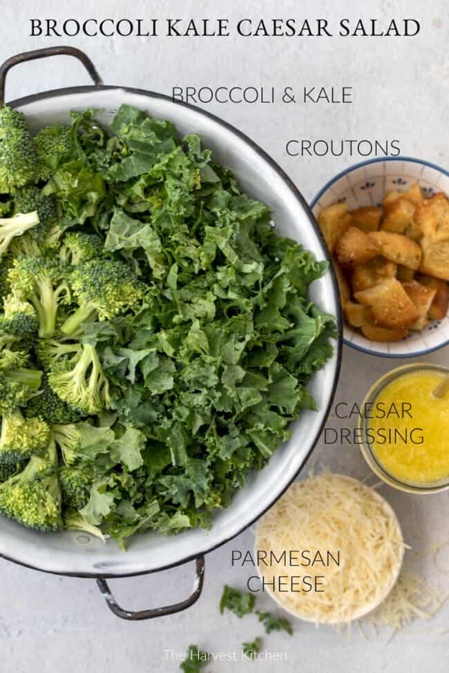 A white colander with black handles filled with raw broccoli and chopped kale. Small bowls sit next to the colander filled with croutons, parmesan cheese and salad dressing.