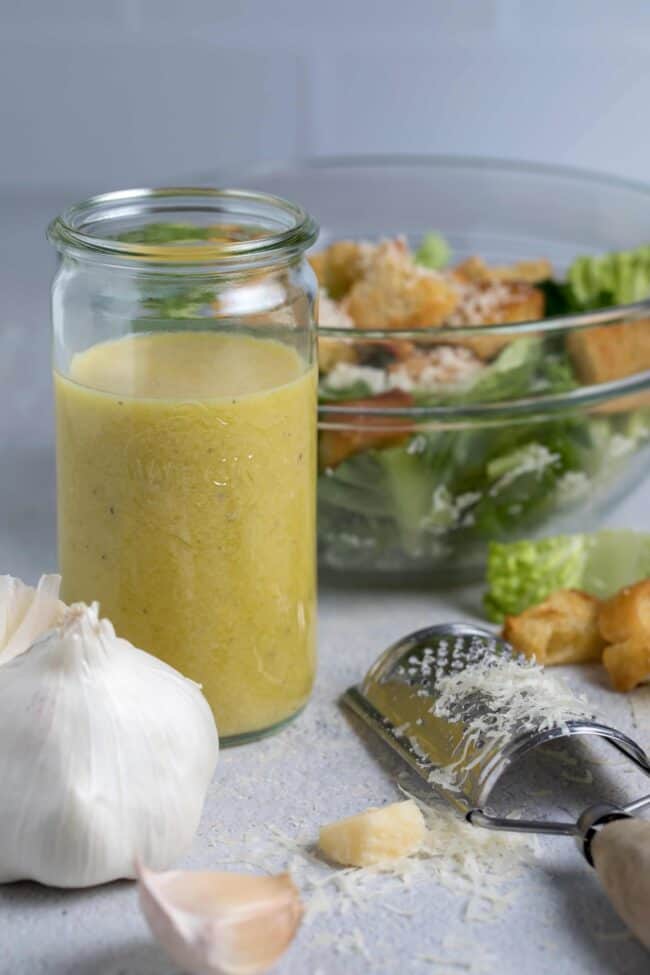 Clear mason jar filled with homemade Caesar dressing. Garlic clove, parmesan cheese and grater next to jar. Glass bowl in background filled with salad.