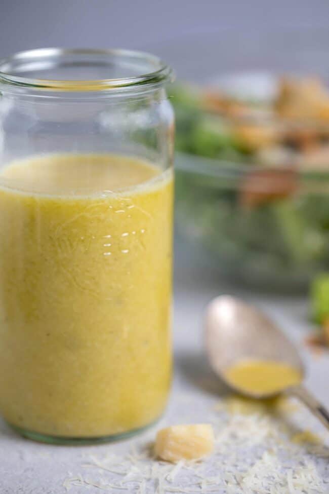 Clear mason jar filled with homemade Caesar dressing. Glass bowl in background filled with salad and spoon lying next to jar