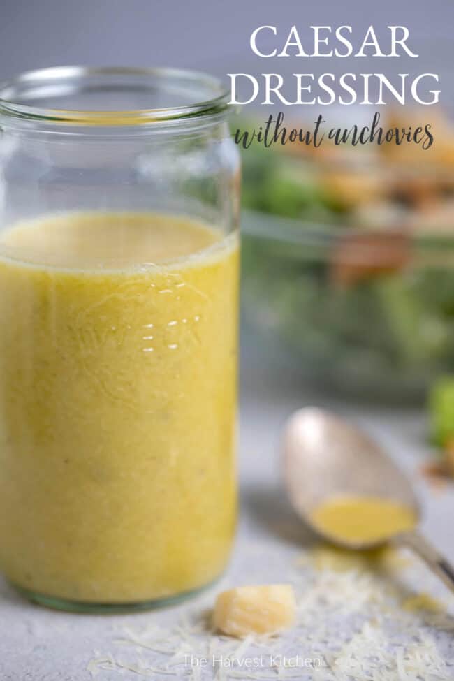 Clear mason jar filled with homemade Caesar dressing. Glass bowl in background filled with salad and spoon lying next to jar.