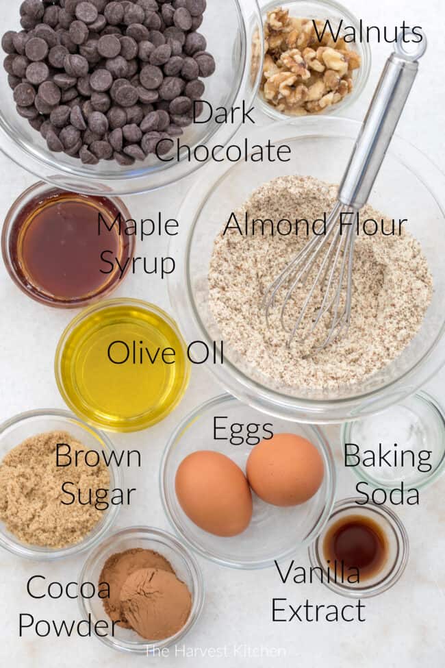 Small glass bowls filled with chocolate chips, sugar, walnuts, oive oil, maple syrup and cocoa powder.